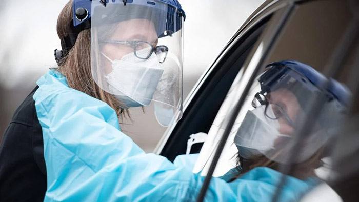 A woman in blue lab coat and face mask looking out of car window.